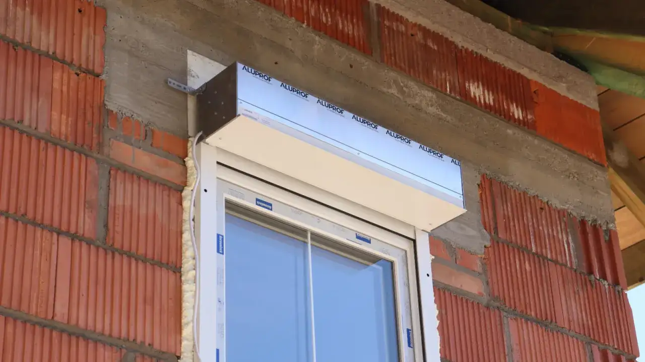 Rozszczelnione okno z widoczną pianką montażową i aluminiową osłoną. Ceglana ściana w trakcie budowy.