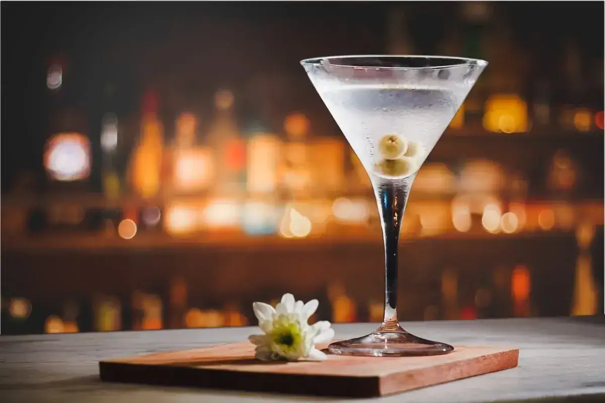Elegancki martini z oliwkami, obok biały kwiatek. Tło rozmyte, barowe. Idealny drink na wieczór.