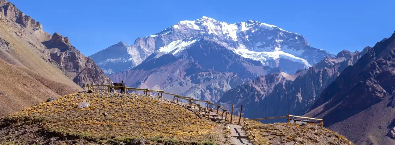 Widok na majestatyczne g&oacute;ry z ośnieżonym Aconcagua w tle. Ścieżka z drewnianym ogrodzeniem prowadzi przez suchą, trawiastą przestrzeń.