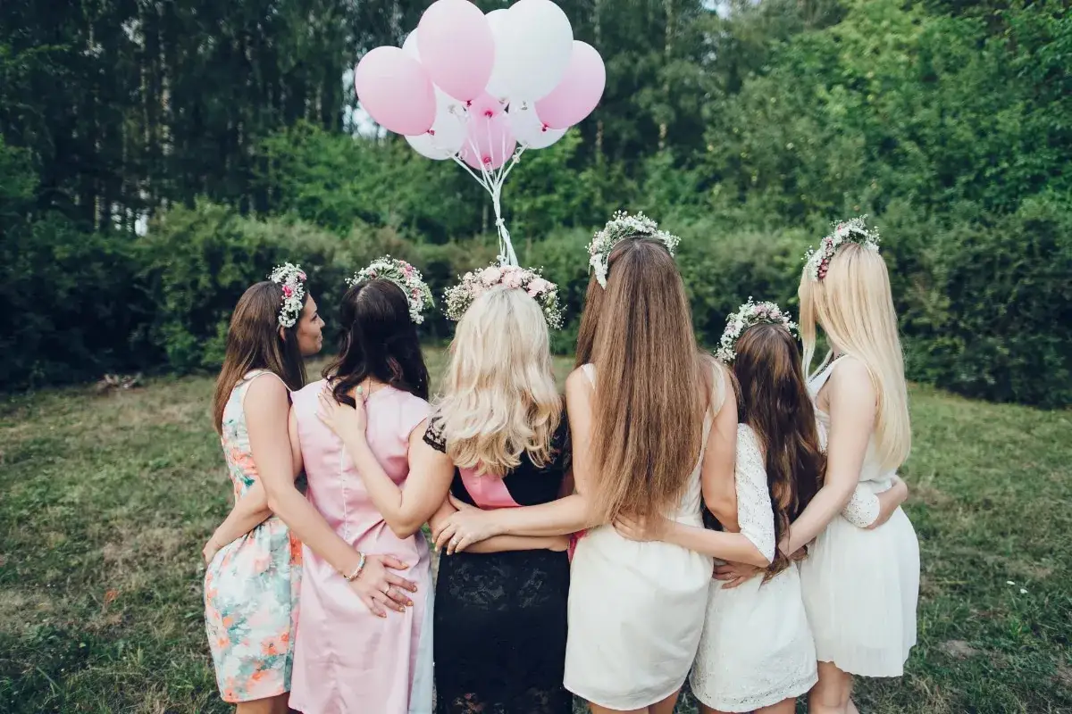 Groupe de femmes en robes d'été et couronnes de fleurs, bras autour des épaules, avec des ballons roses et blancs. Idéal pour des jeux EVJF à imprimer gratuit.