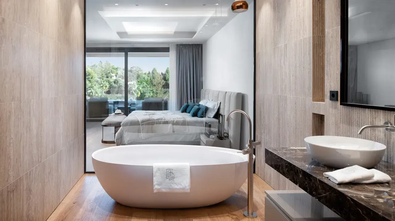 Un lujoso baño con bañera exenta y lavabo sobre encimera de mármol. A través de un cristal, se ve un dormitorio moderno con cama y vistas a un exuberante jardín. ¡Se puede hacer un baño en una habitación así!