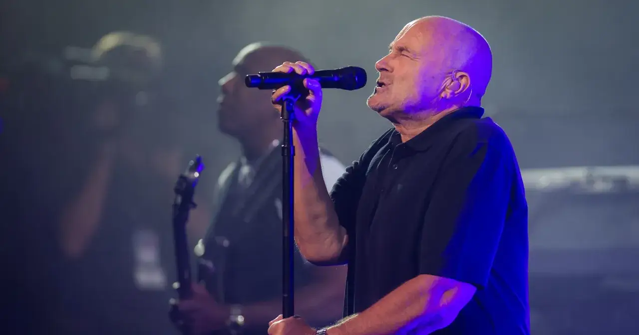 Phil Collins śpiewa na scenie, w tle widać gitarzystę. Choć jego żony nie ma na zdjęciu, jego muzyka porusza serca fanów.
