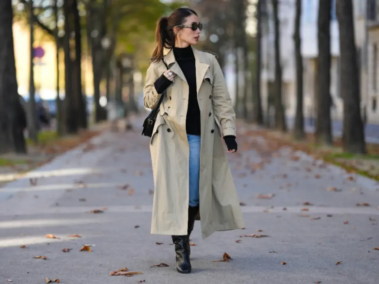Mujer pasea con un elegante trench beige, suéter negro y jeans, luciendo lo que se lleva este otoño.