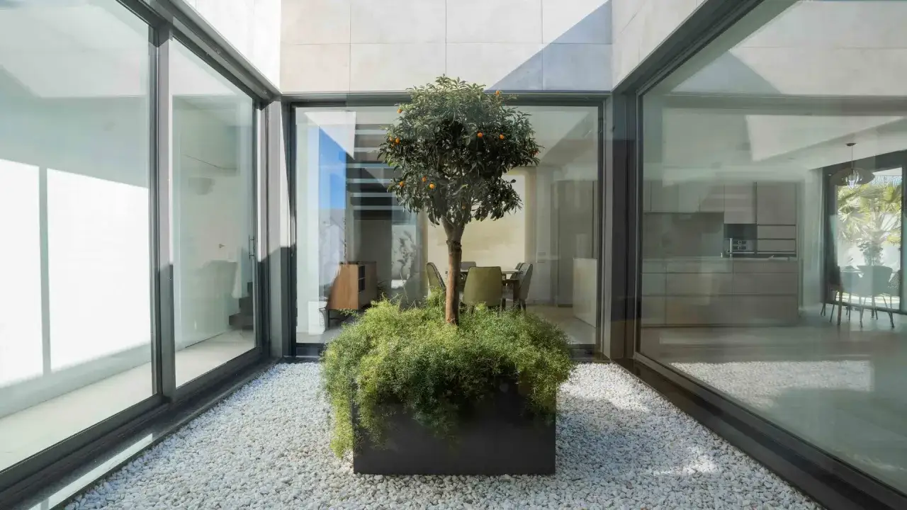 Un &aacute;rbol frutal en un gran macetero preside estos patios interiores con encanto, rodeado de grava blanca y cristaleras que conectan con el comedor y la cocina.