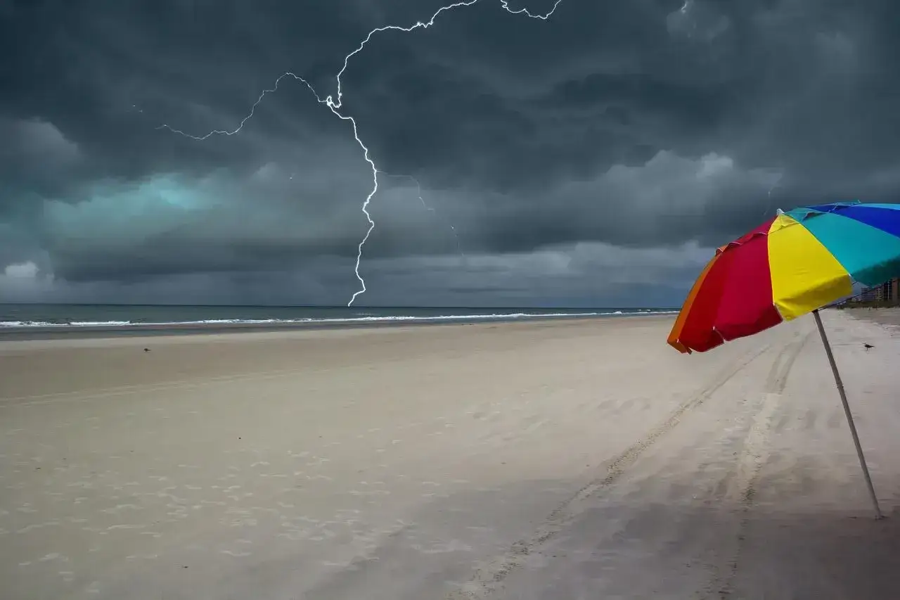 Plaża pustoszeje, gdy nadciąga gwałtowny **szkwał**. Błyskawica przecina ciemne chmury, a kolorowy parasol stoi samotnie na piasku.