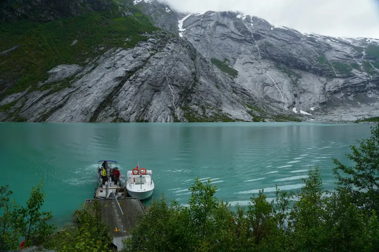 Turkusowa woda i strome góry to obraz tego, co to są fiordy w Norwegii. Dwa statki czekają przy molo.