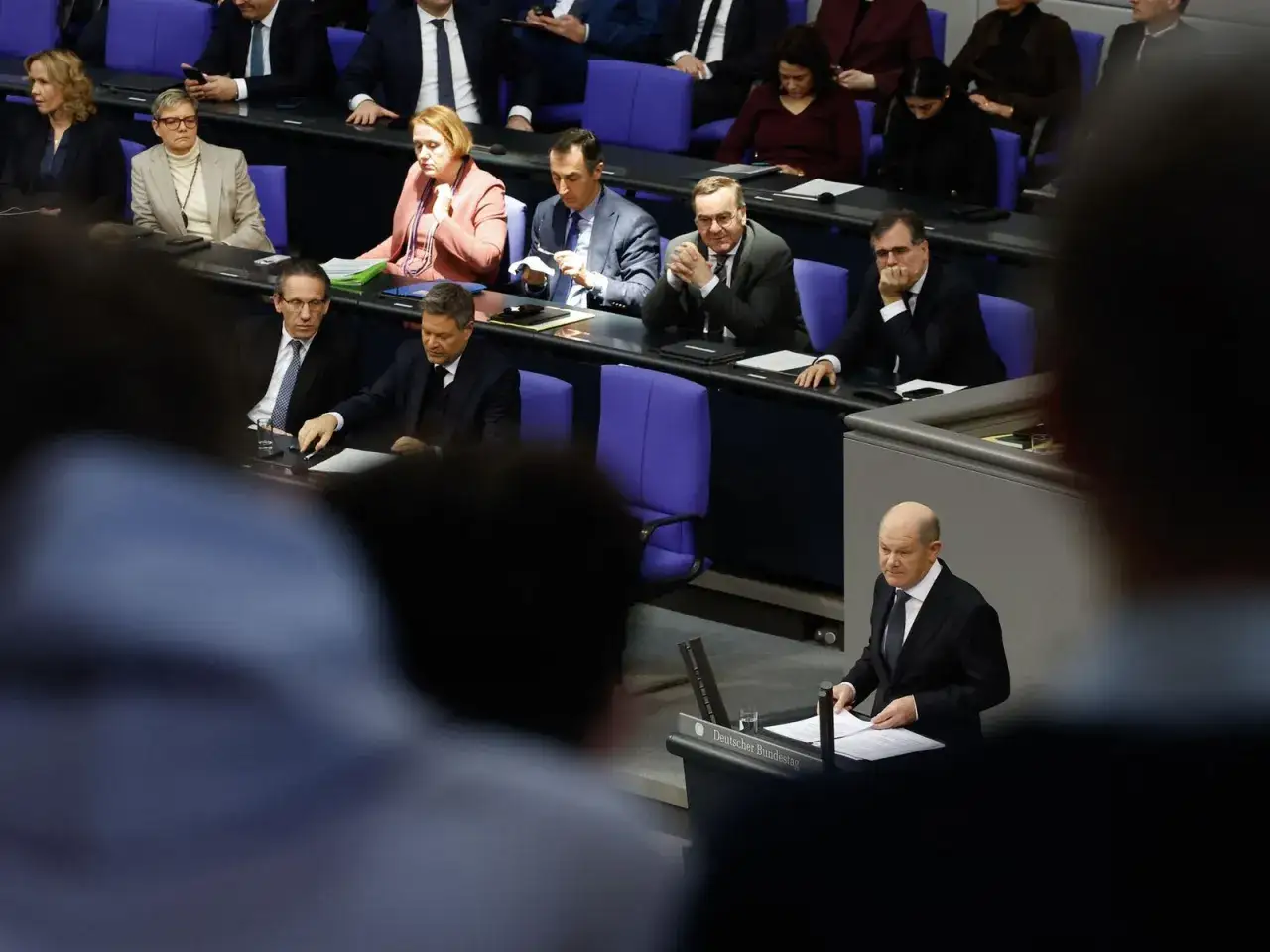 Olaf Scholz spricht im Bundestag. Die Gesichter der Abgeordneten zeigen unterschiedliche Reaktionen, was die polarisierende Politik widerspiegelt.