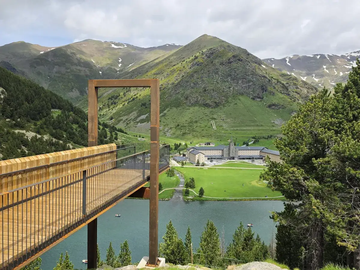 Mirador moderno sobre el lago en Vall de Núria, ideal para explorar las **rutas Vall de Nuria**. Montañas verdes y un santuario al fondo.