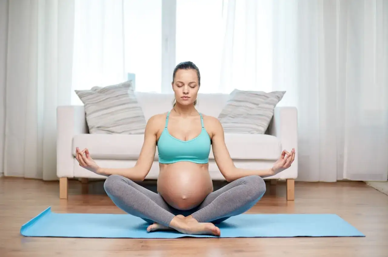 Schwangere Frau in Lotussitz auf Yogamatte, Hände im Mudra, Augen geschlossen. Entspannende Meditation für Schwangerschaft.