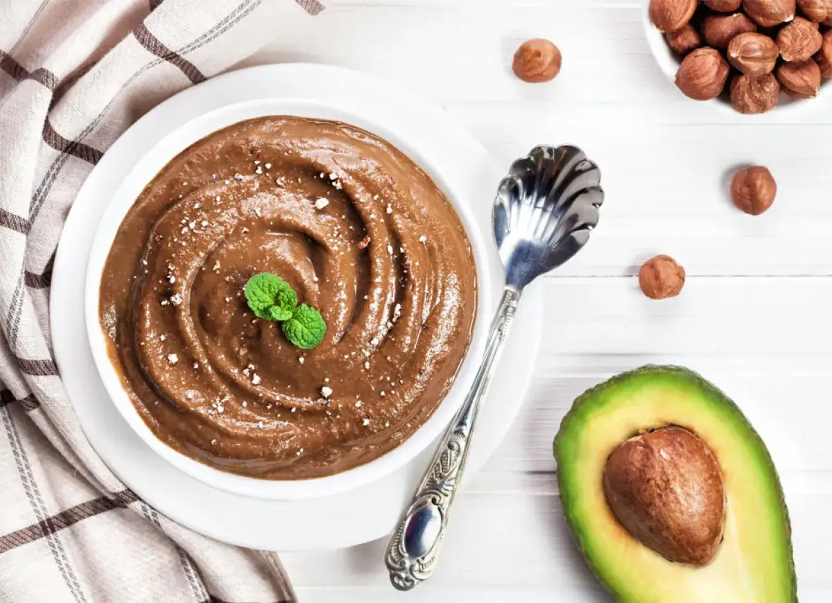 Kremowa nutella z awokado, udekorowana miętą, obok orzechów laskowych i połówki awokado.