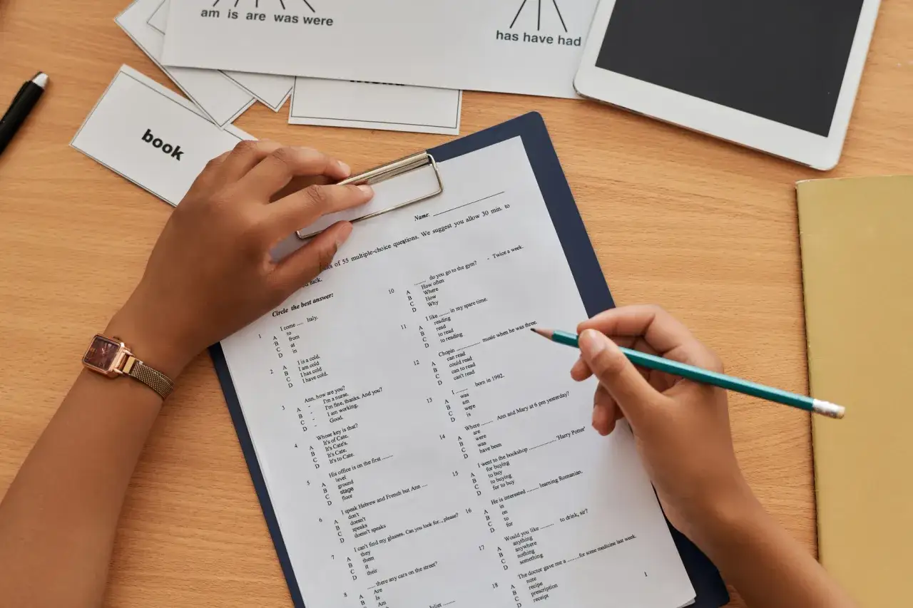 Ręce ucznia piszą test. Co jeśli nauczyciel zgubi prace ucznia? Na biurku leżą karty z angielskimi słówkami i tablet.