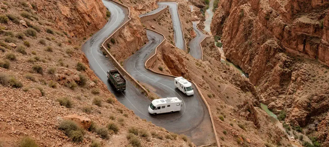 Deux camping-cars naviguent sur une route sinueuse &agrave; flanc de montagne, offrant un aper&ccedil;u du Maroc en camping-car. Un camion traverse la sc&egrave;ne.