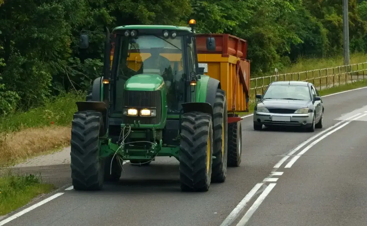 Zielony traktor z przyczepą jedzie po drodze. Obok niego jedzie samochód, a linia podwójna ciągła sugeruje, że nie można wyprzedzić traktora.