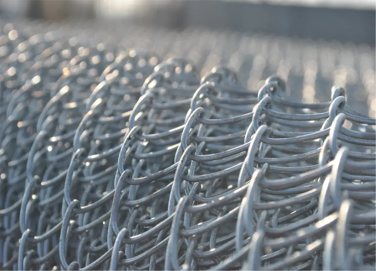 Zbliżenie na zwinięte ogrodzenie z siatki ocynkowanej. Metalowe ogniwa tworzą regularny wz&oacute;r, odbijając światło.