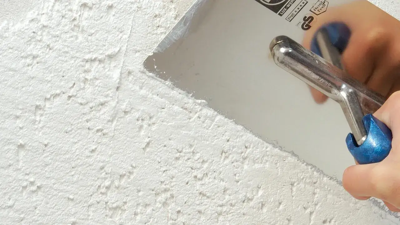 Hand mit Gl&auml;ttekelle bearbeitet eine wei&szlig;e, strukturierte Wand. Das Werkzeug verteilt den Putz, um die Oberfl&auml;che f&uuml;r das Fliesen verputzen vorzubereiten.