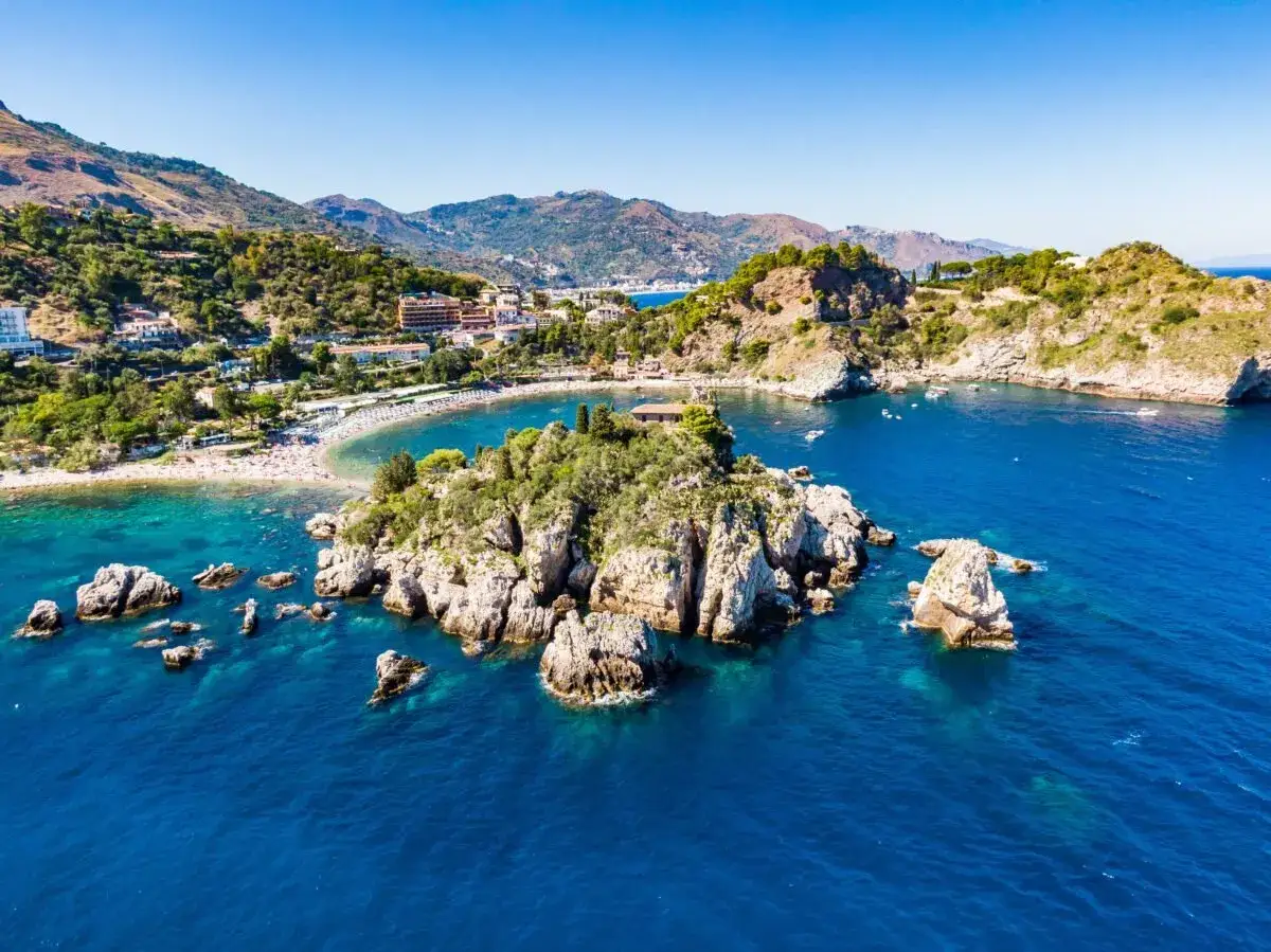 &Icirc;le rocheuse verdoyante baign&eacute;e par les eaux bleues de la mer, avec une plage anim&eacute;e et des collines en arri&egrave;re-plan, pr&egrave;s de Catane, Sicile.