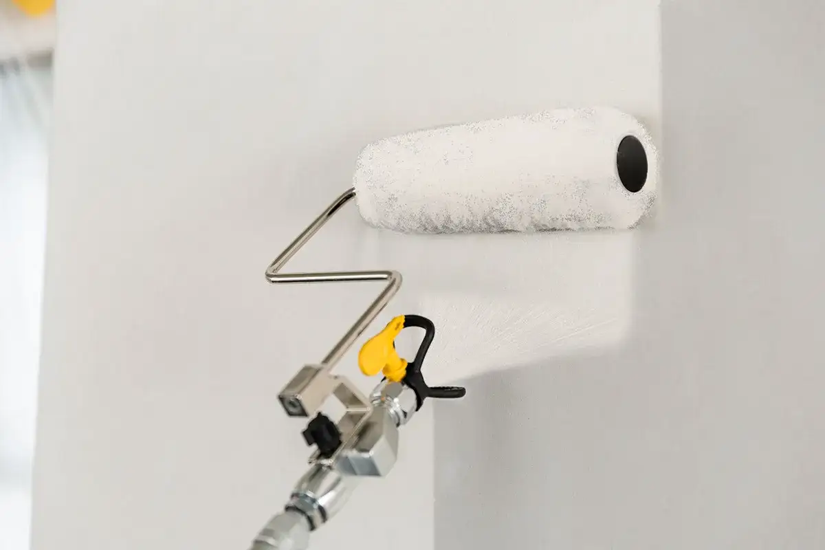 Fasadm&aring;lning med roller och sprutpistol. En vit roller applicerar f&auml;rg p&aring; en ljus v&auml;gg.