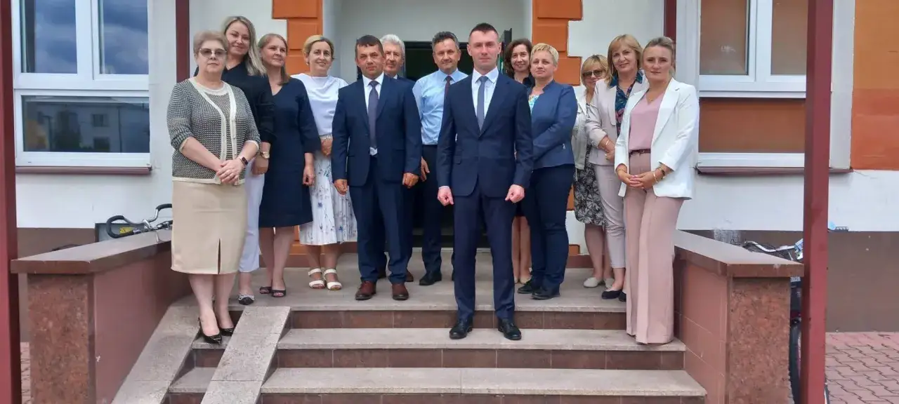 Grupa osób pozuje przed budynkiem szkoły. Wszyscy ubrani są elegancko, co sugeruje, że uczestniczą w ważnym wydarzeniu, być może w konkursie na dyrektora szkoły.