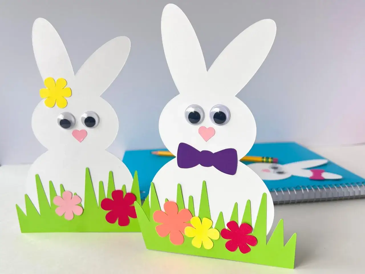 Zwei süße Osterhasen aus Papier, die Kinder leicht basteln können, stehen im grünen Gras mit bunten Blumen.