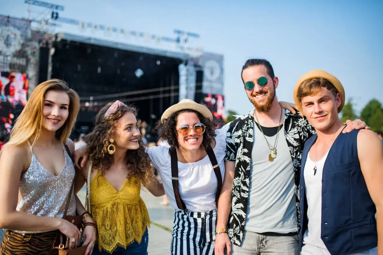 Piątka przyjaciół w letnich stylizacjach, idealnych na koncert. Wszyscy uśmiechnięci, w tle scena festiwalowa.