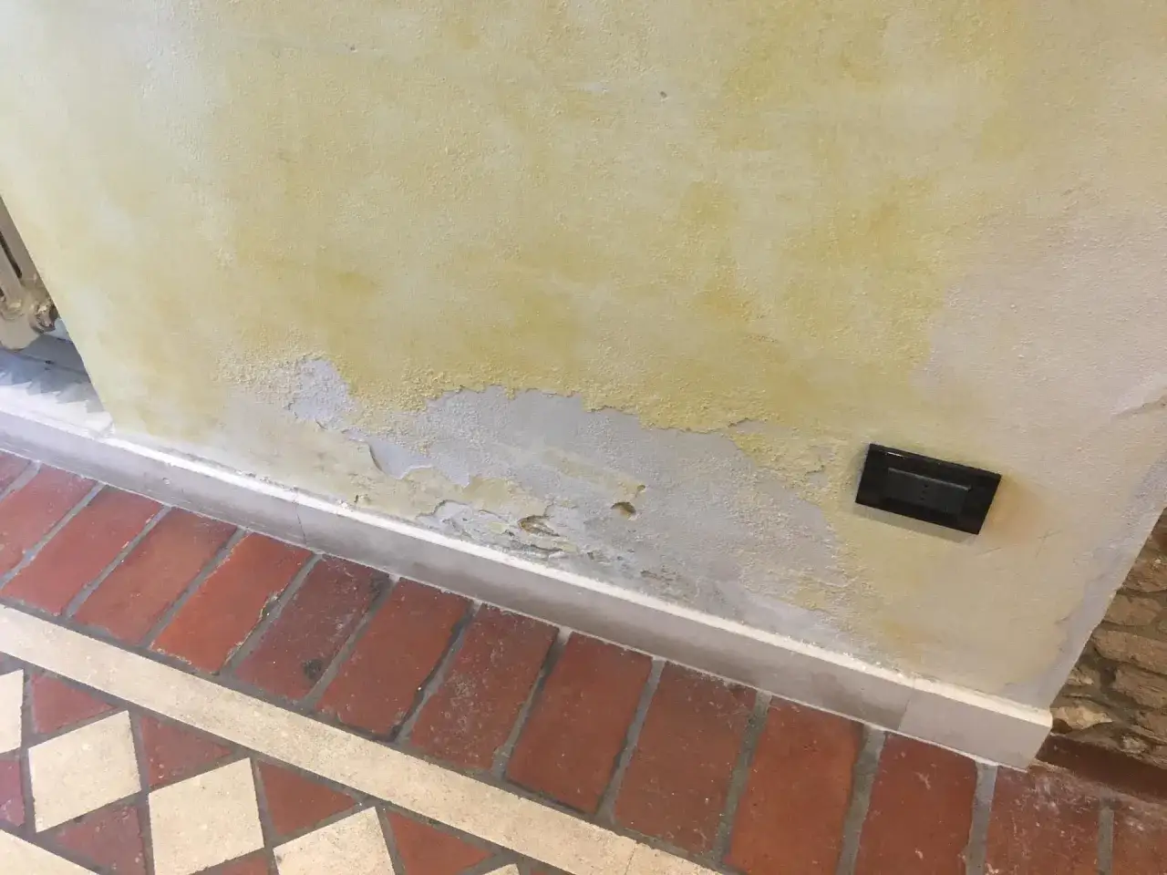 Muro ingiallito e scrostato al piano terra, segno di umidità di risalita. Pavimento in cotto e inserti decorativi.