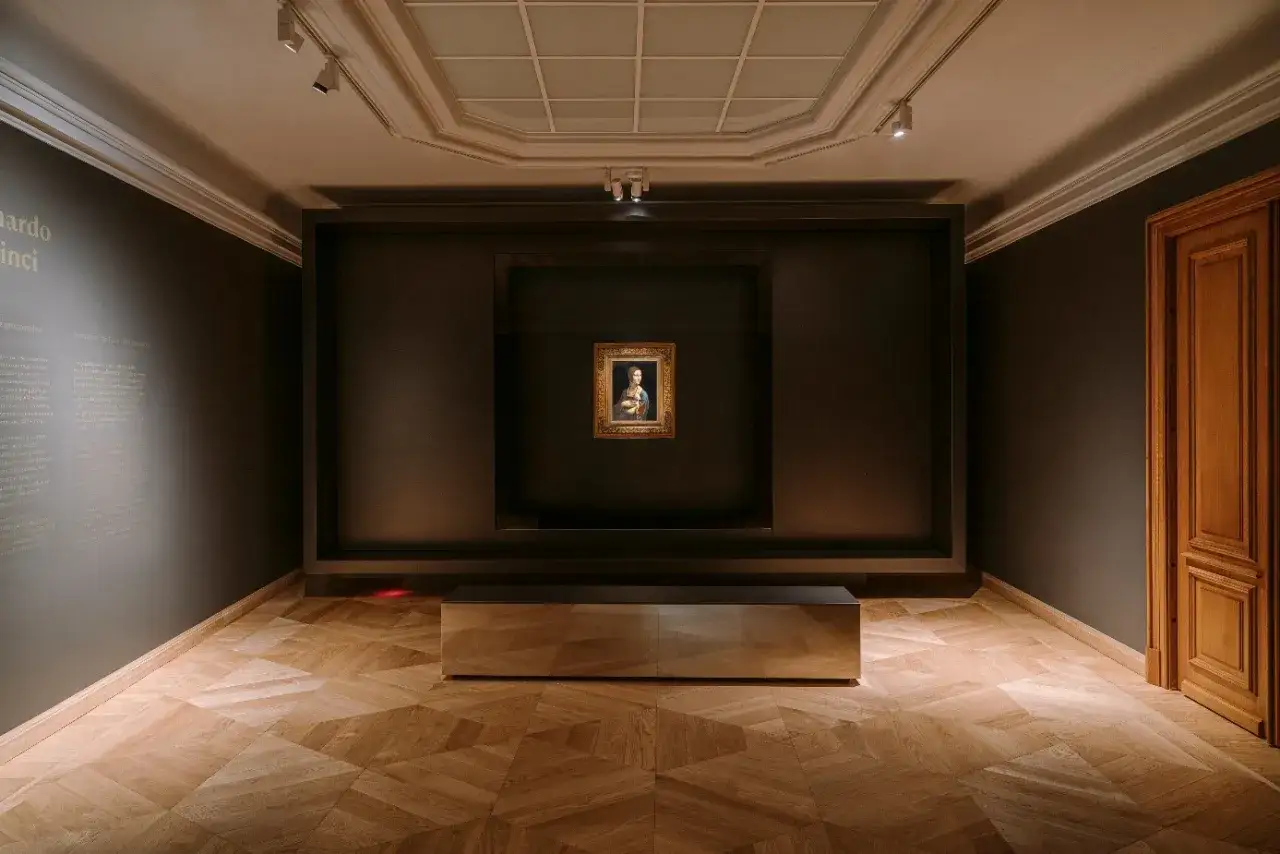 Dama z łasiczką Leonarda da Vinci w muzeum, gdzie jest? Obraz w złotej ramie na ciemnej ścianie, pod nim ławka.