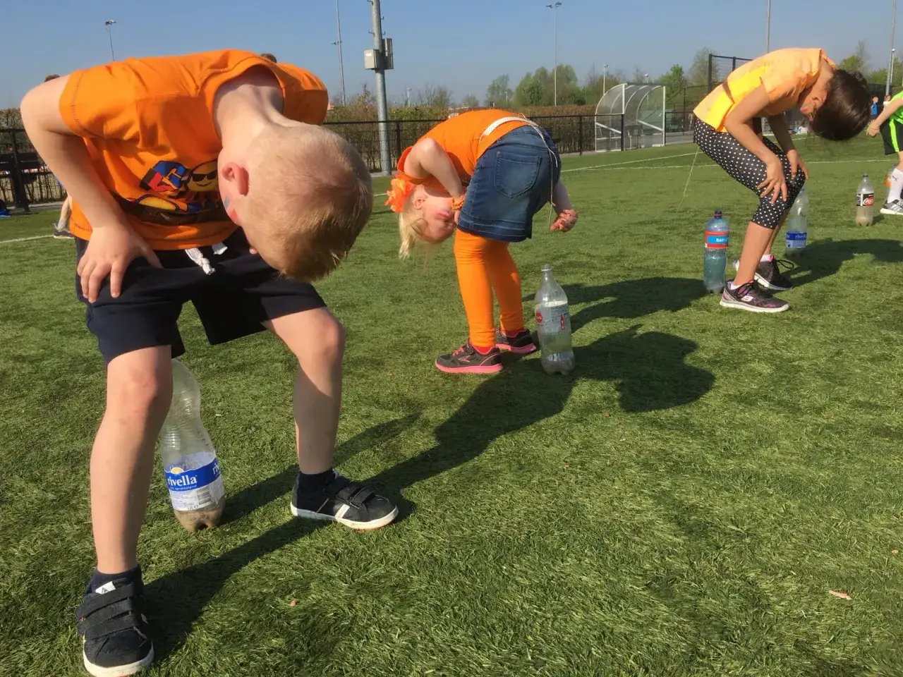 Barn i orange tr&ouml;jor deltar i lekar utomhus p&aring; en gr&auml;splan, b&ouml;jda &ouml;ver flaskor.