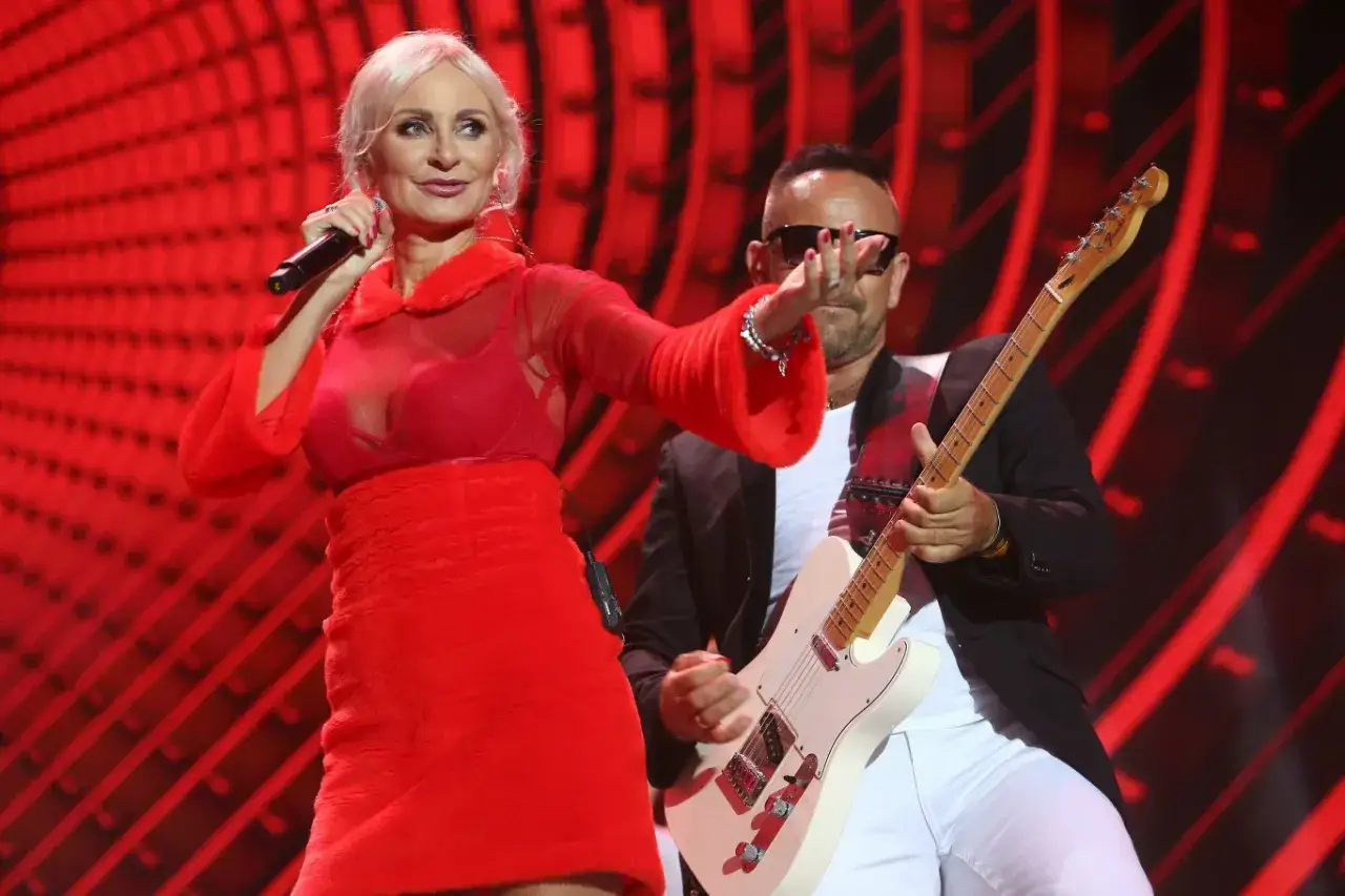 Elwira Mejka, w czerwonej sukience, śpiewa na scenie. Obok niej gitarzysta.