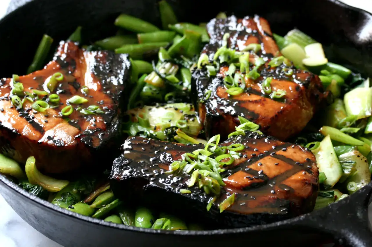 Three thick, glazed miso pork chops rest in a cast iron skillet with vibrant green beans and bok choy, garnished with fresh scallions and black sesame seeds.