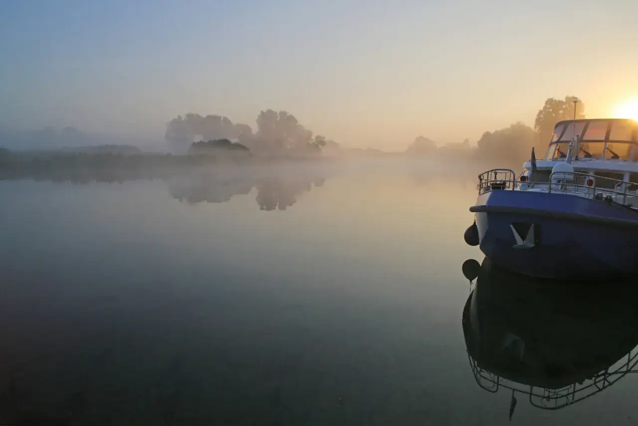 Ein blaues Schiff liegt still im Nebel auf einem ruhigen Fluss. Die aufgehende Sonne wirft einen warmen Schein auf die Szene.