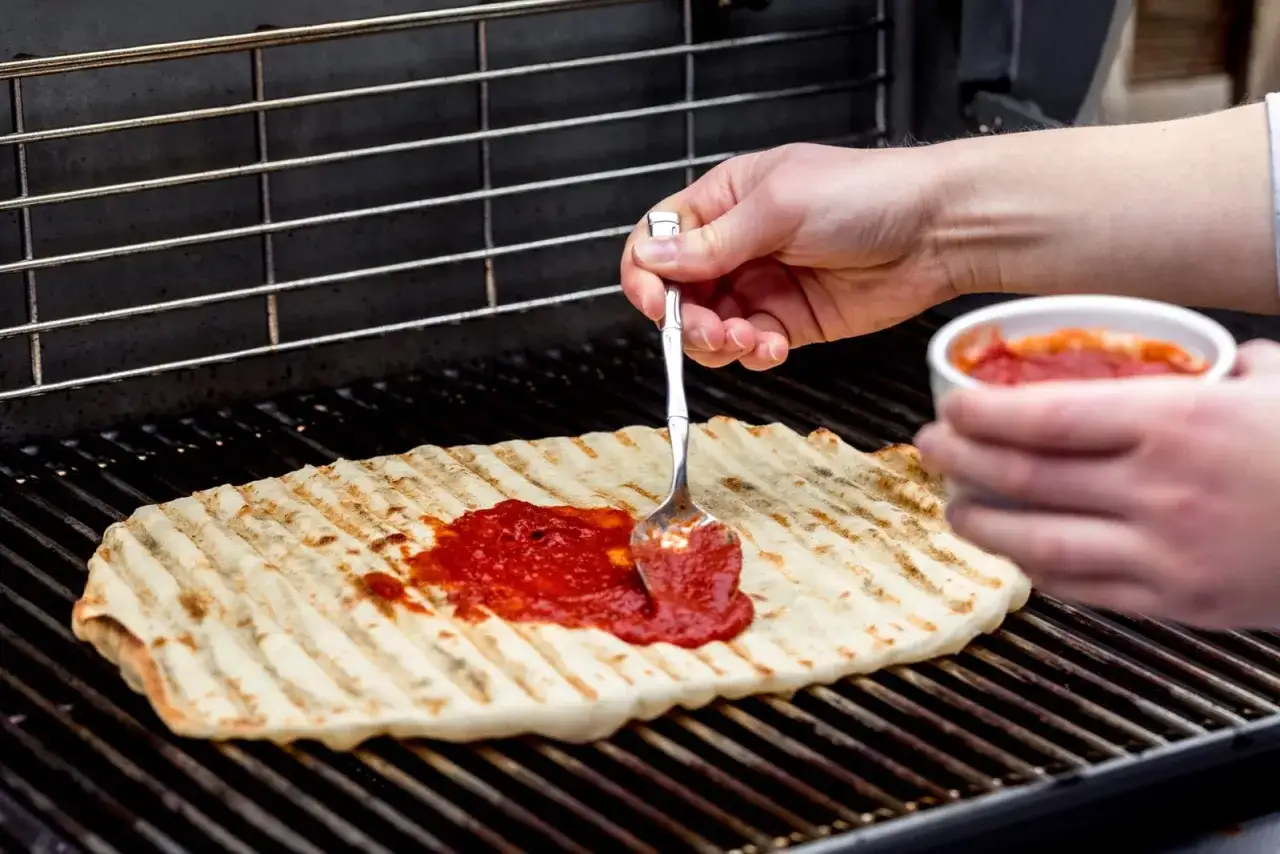 En person sprider tomats&aring;s p&aring; en grillad pizzabotten p&aring; en grill.