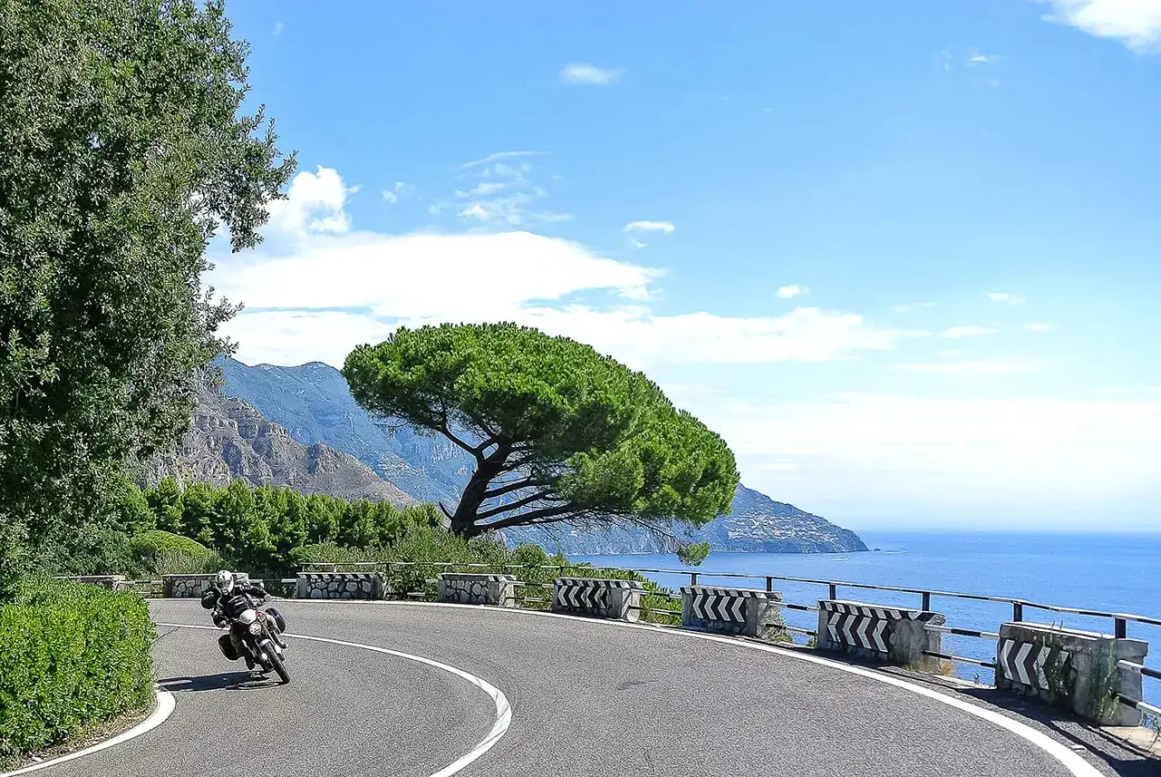Motociclista percorre le curve delle **strade costiera amalfitana**, con il mare blu e montagne sullo sfondo.