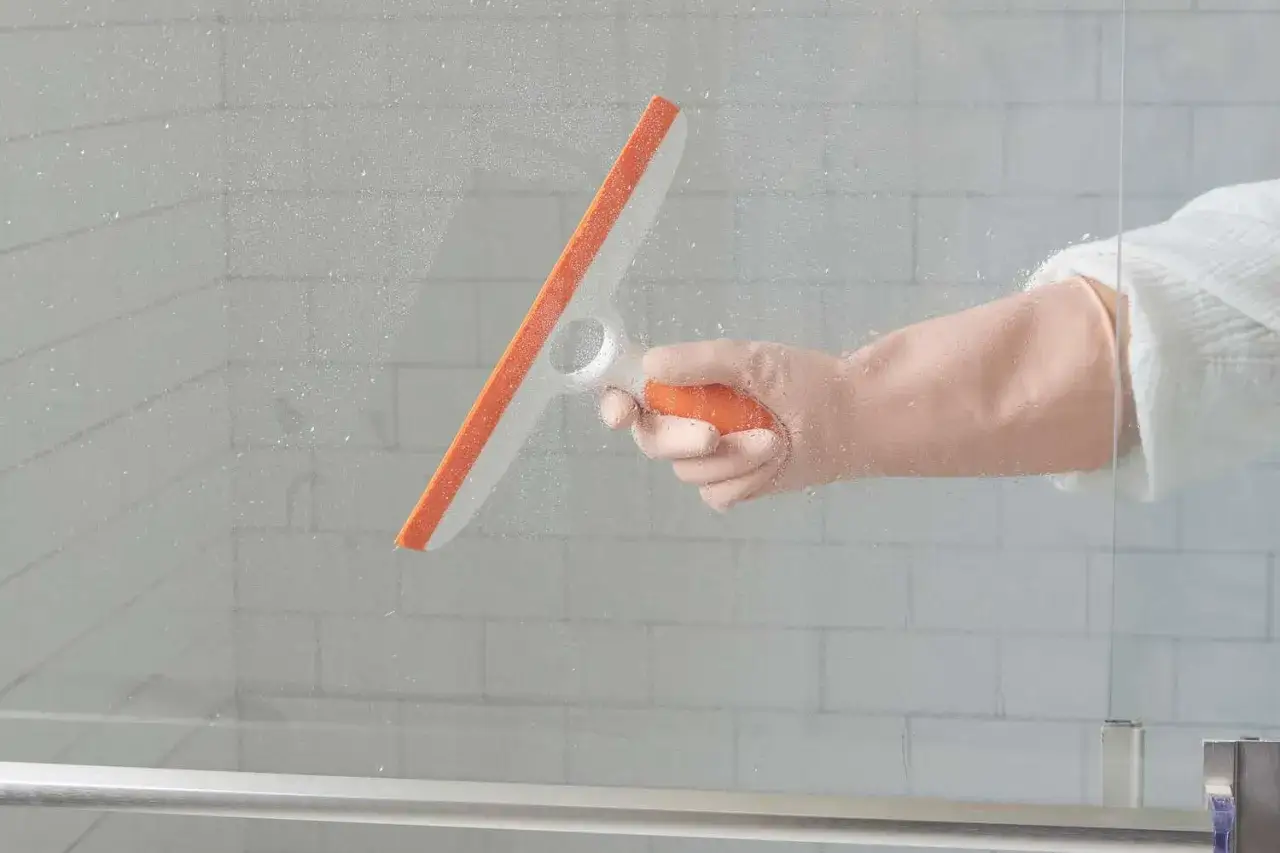 Main tenant, ganté, utilise un racloir pour nettoyer vitre douche.