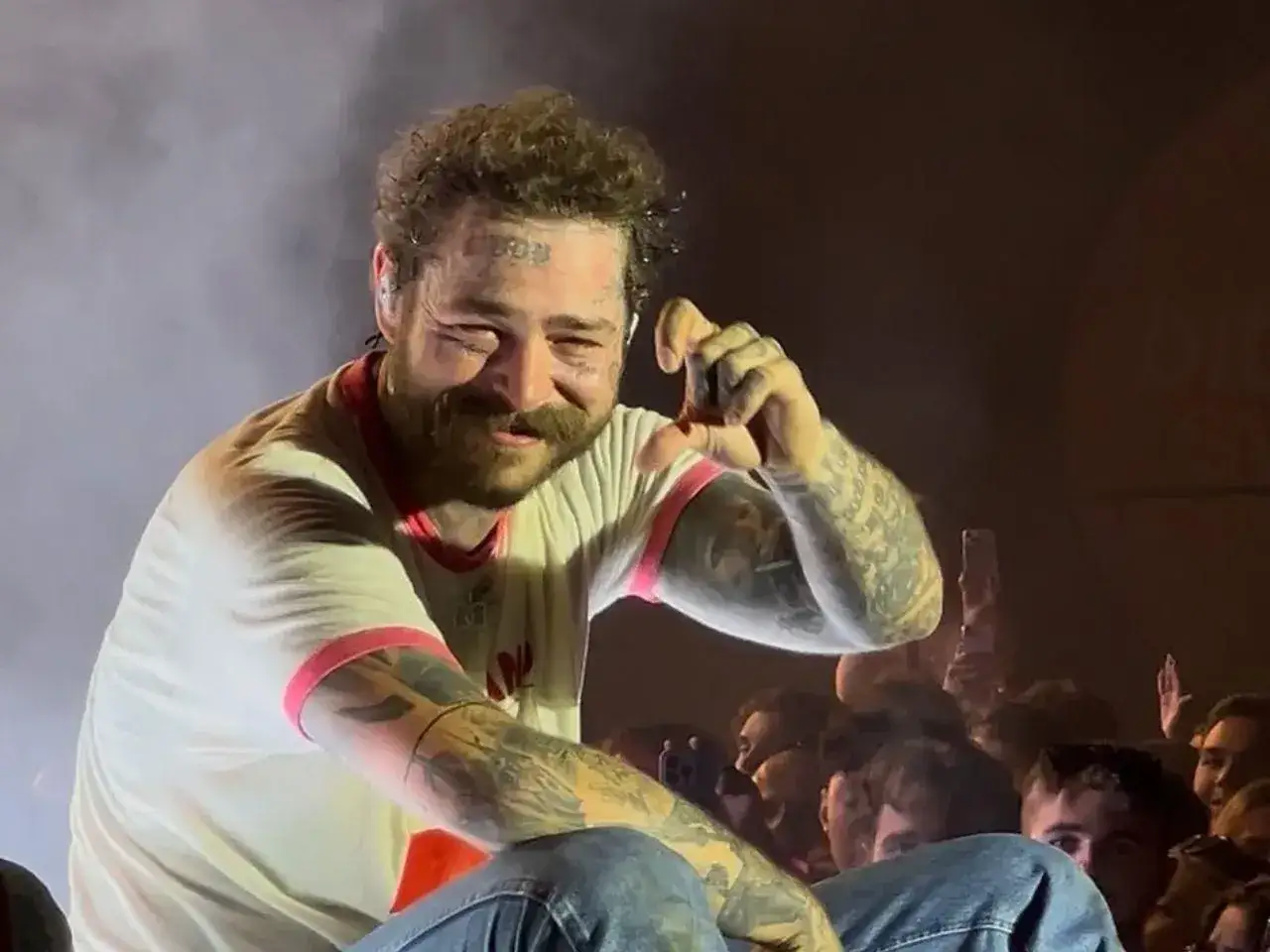 Post Malone Polska na koncercie, uśmiechnięty, z tatuażami na twarzy i rękach, pozdrawia fanów.