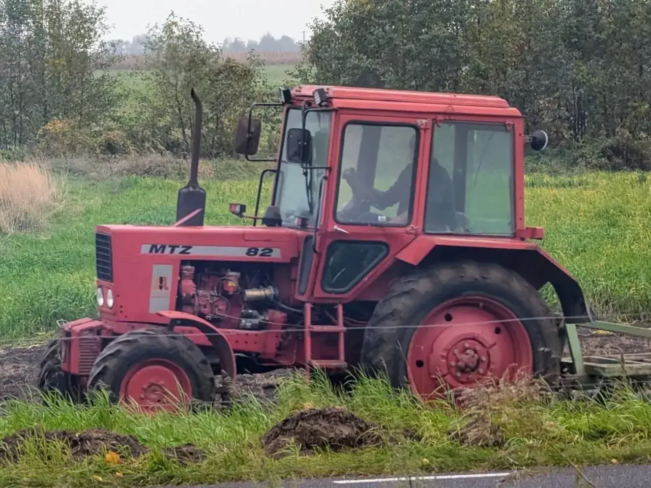 Czerwony traktor Belarus 820 pracuje na polu, za nim widać las i zielone pola.