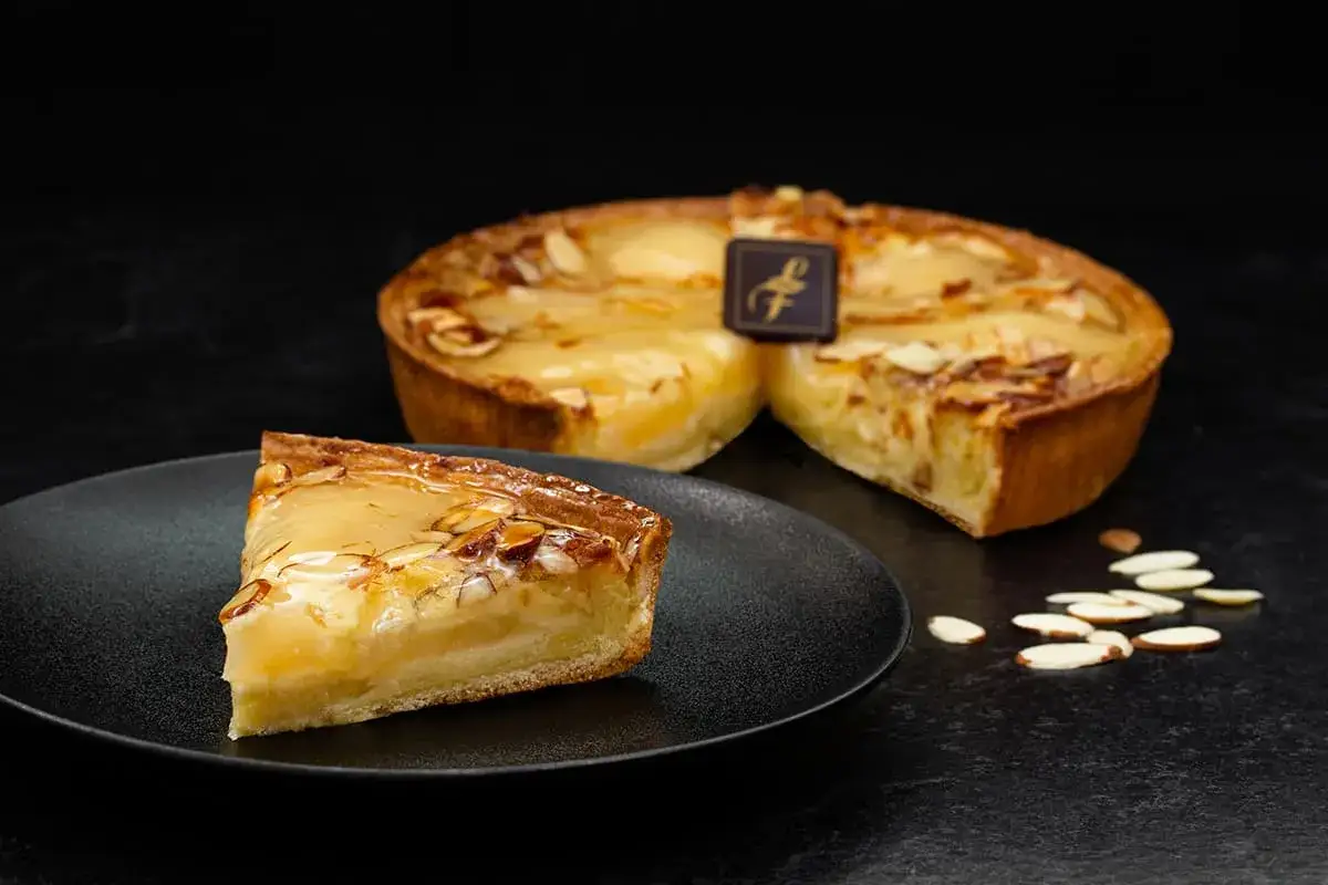 Une part de tarte aux poires, garnie d'amandes effil&eacute;es et d'un gla&ccedil;age brillant, est pos&eacute;e sur une assiette noire. Une tarte enti&egrave;re, d&eacute;cor&eacute;e d'un logo, est visible en arri&egrave;re-plan.