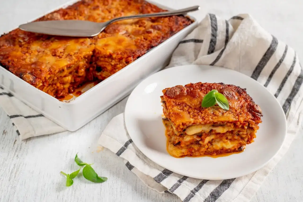 Una porzione di melanzane alla parmigiana su un piatto bianco, con la teglia ancora piena sullo sfondo.