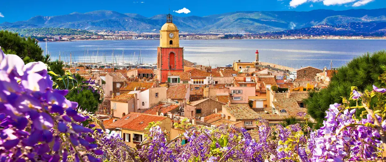 Blick auf das charmante St. Tropez mit seinem markanten Kirchturm, roten Ziegeldächern und dem glitzernden Meer im Hintergrund, umrahmt von lila Blüten.