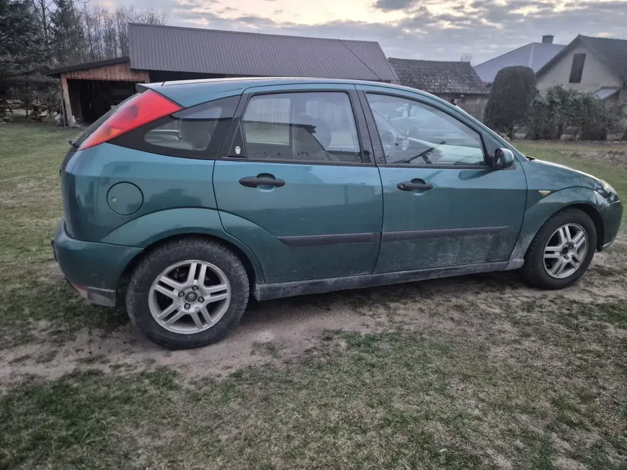 Zielony Ford Focus MK1 1.6 benzyna, zaparkowany na trawiastym podwórku, z widocznymi budynkami w tle.