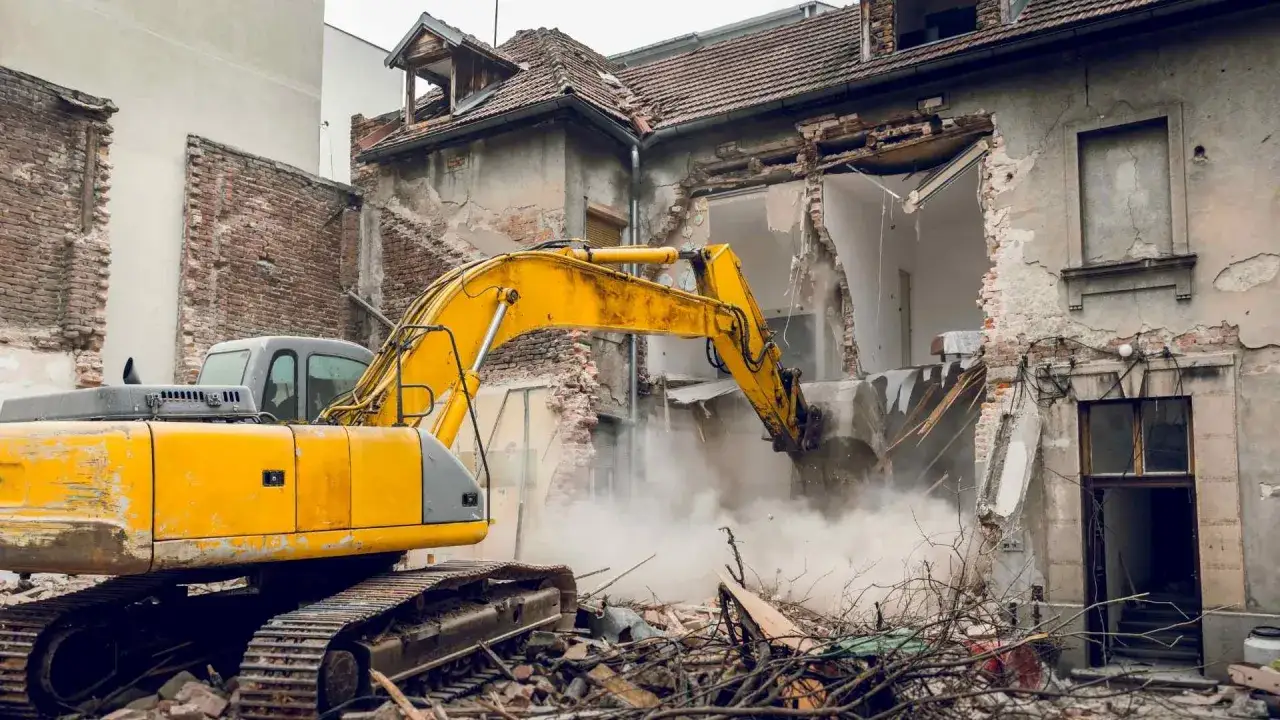 Une pelleteuse jaune démolit une maison en briques, soulevant un nuage de poussière. Le chantier donne une idée du prix démolition maison.