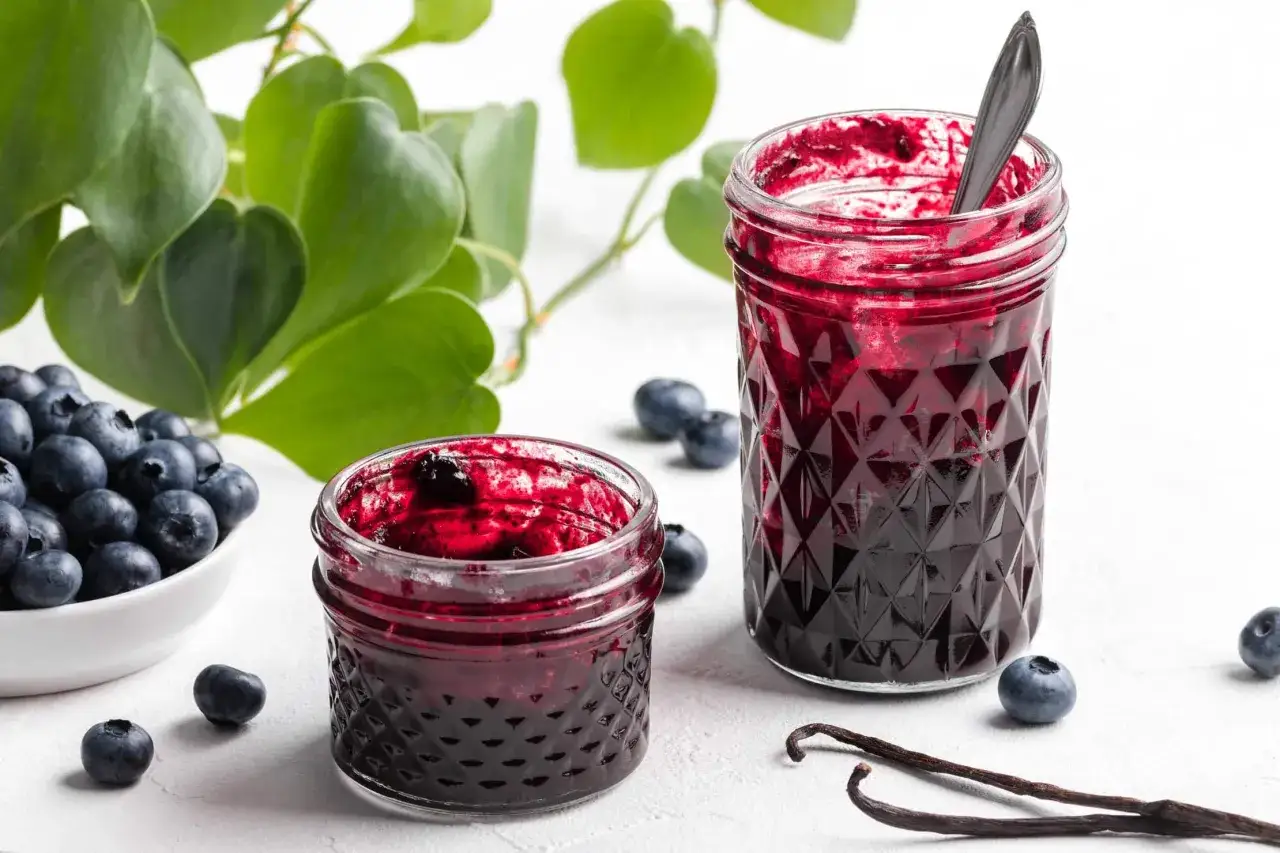 Zwei Gläser mit ausgefallener Heidelbeermarmelade, daneben frische Beeren und Vanilleschoten.