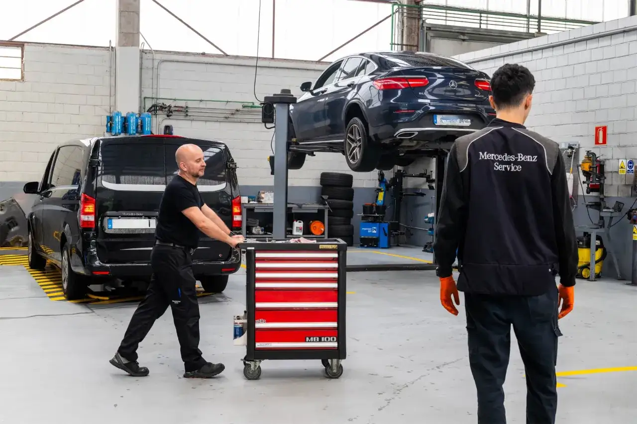 Taller mecánico Mercedes-Benz. Un técnico empuja un carrito de herramientas mientras otro, con uniforme de servicio, supervisa el mantenimiento de un Mercedes-Benz GLC elevado.