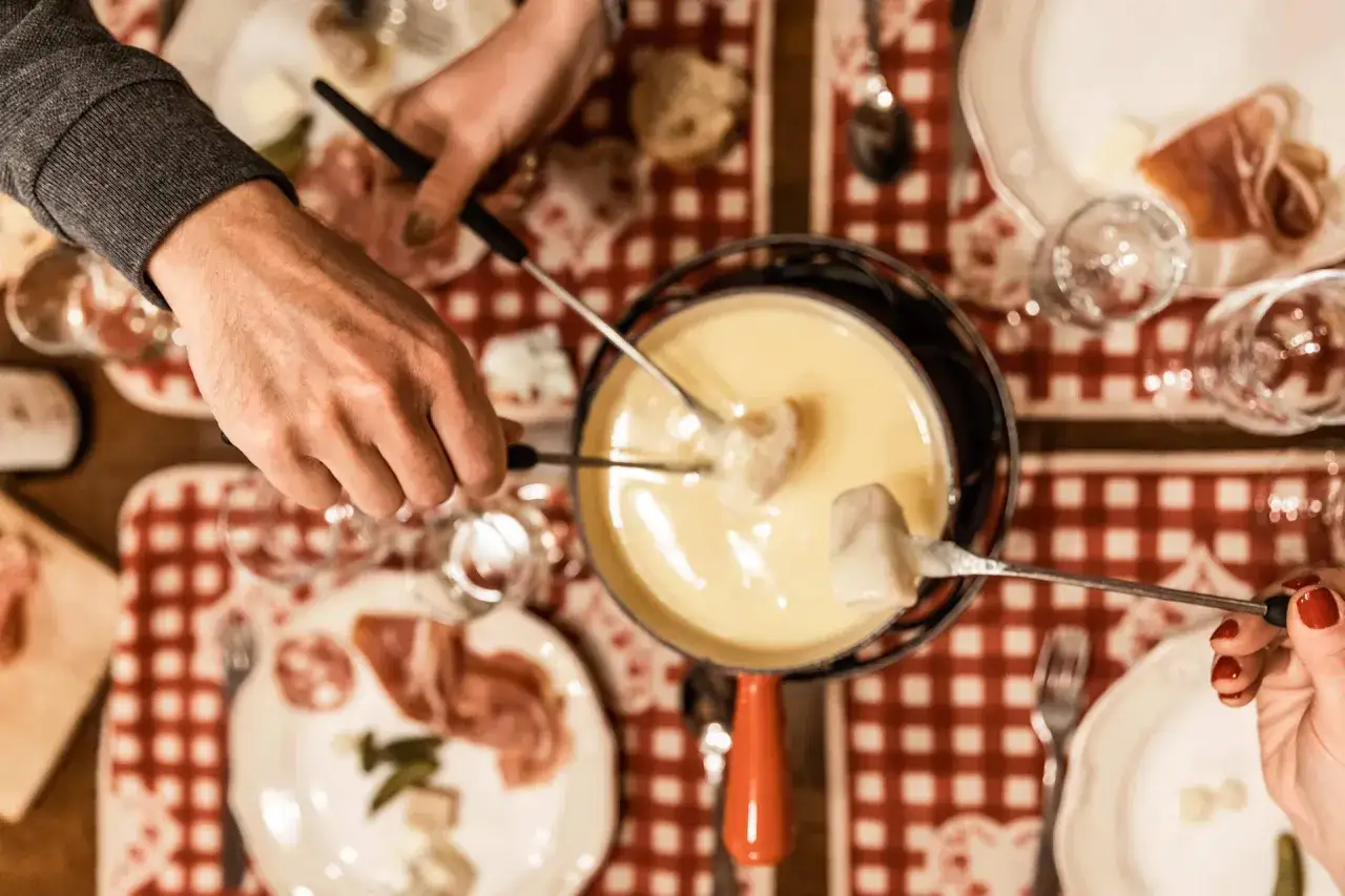 Un convivial repas fromage fondu partagé entre amis. Des brochettes trempent dans le caquelon, entourées de charcuterie et de pain.