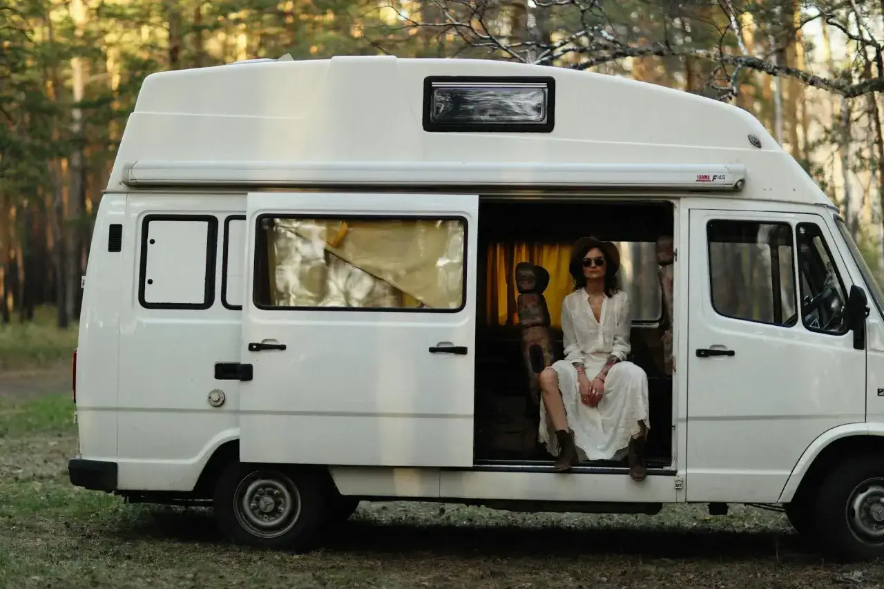 Kobieta w białej sukience i kapeluszu siedzi w otwartych drzwiach białego kampera, uosabiając wolność polskiego caravaningu.