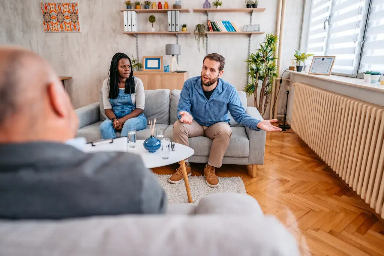 Een man en vrouw, duidelijk conflict zoekende mensen, zitten tegenover een therapeut. De man gebaart met zijn handen, de vrouw kijkt weg.