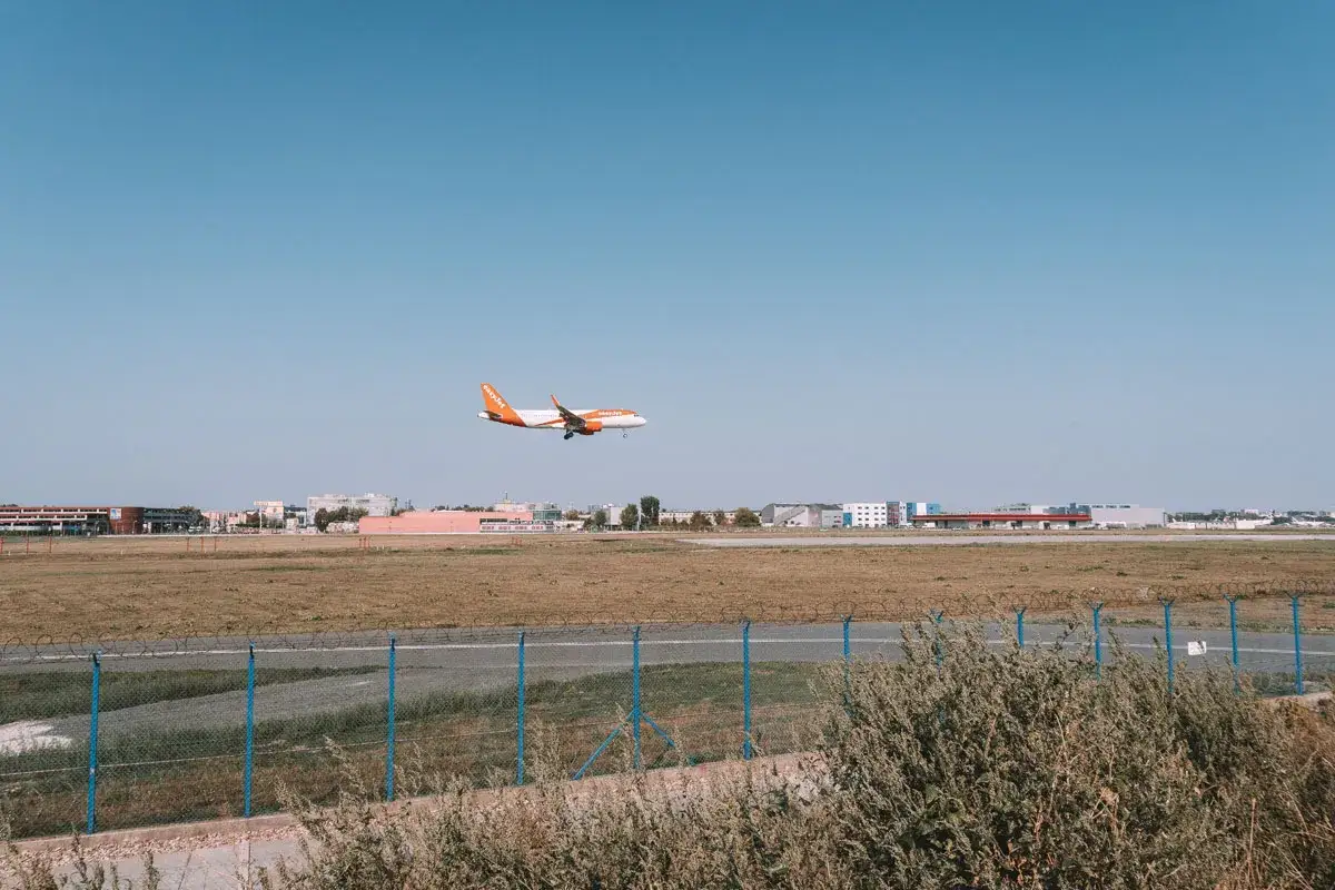 Widok z punktu widokowego lotnisko. Pomarańczowy samolot easyJet zbliża się do lądowania na tle błękitnego nieba i budynk&oacute;w lotniska.
