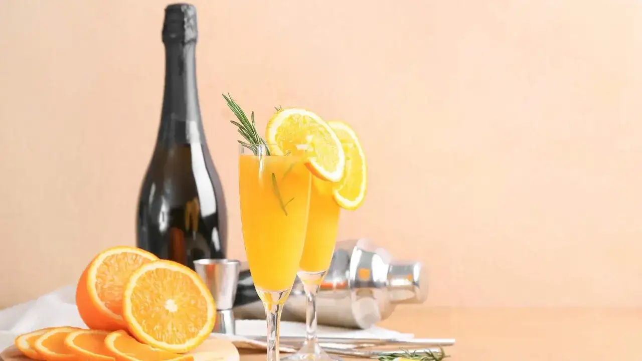 Dwa kieliszki orzeźwiającej mimosa z plasterkami pomarańczy i gałązką rozmarynu, obok butelki szampana i shakera.