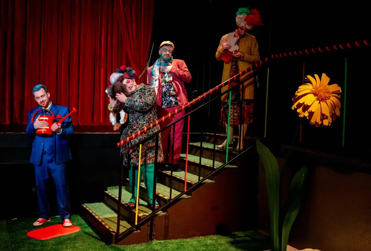 Aktorzy w barwnych strojach i perukach biorą udział w sztuce teatralnej po angielsku. Na scenie duży, żółty kwiat i czerwone zasłony.