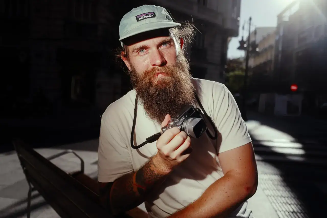 Homme barbu avec casquette Patagonia tenant un appareil photo Leica Q3 pour un test. Lumière du soleil sur son visage.