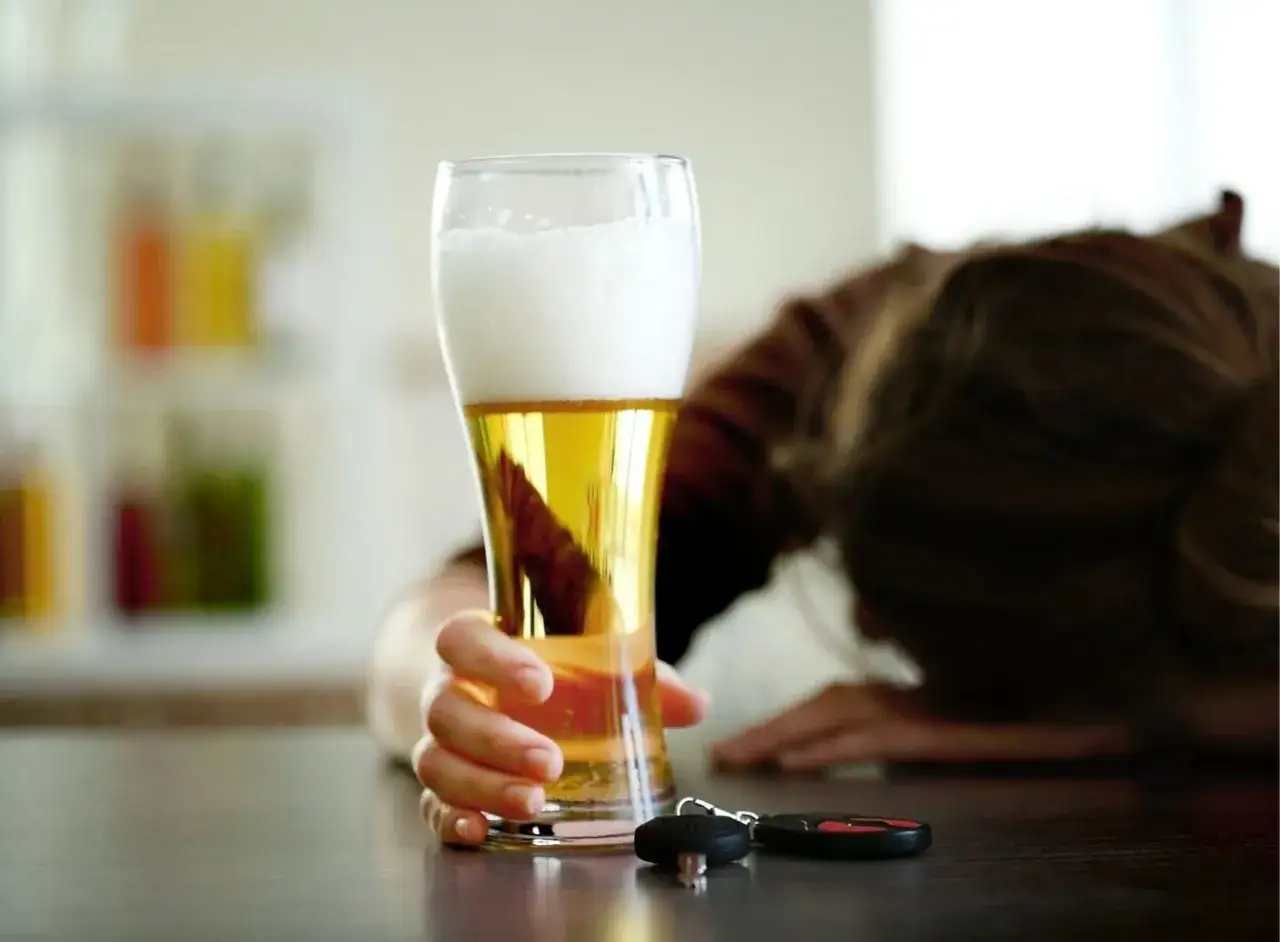 Ręka sięga po jedno piwo, obok leżą kluczyki do samochodu. Osoba w tle jest wyraźnie zmęczona lub pijana.